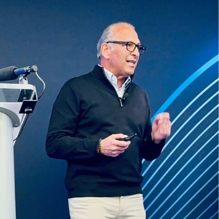 Man in glasses and black sweater gesturing while giving a presentation against blue background.